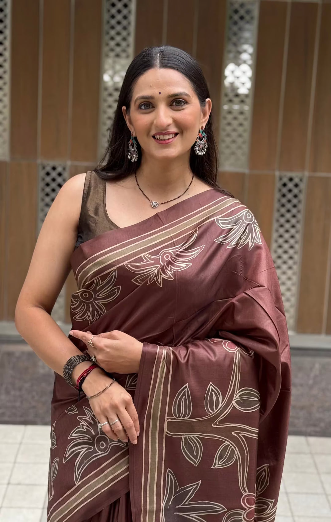 A woman wearing a brown silk saree with beige floral patterns, standing in front of a tiled wall.

