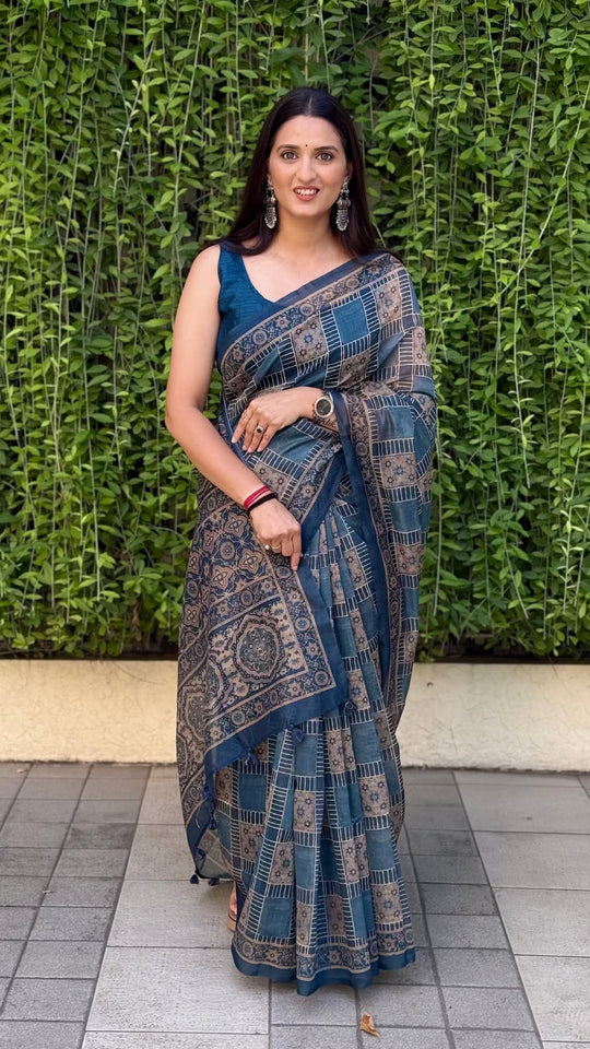 Woman in slate blue color saree stading against the green leafy wall.