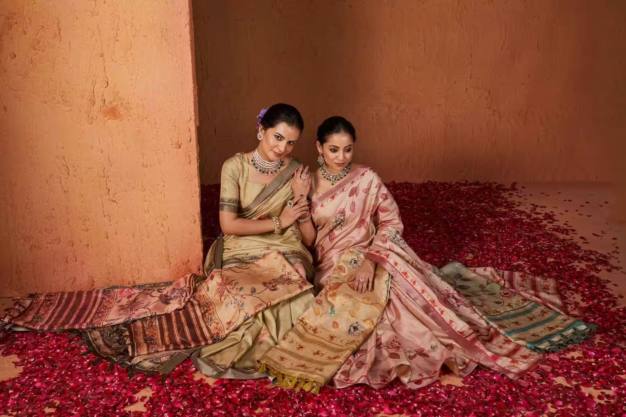 Two women in printed sarees sitting on a floor covered with rose petals.