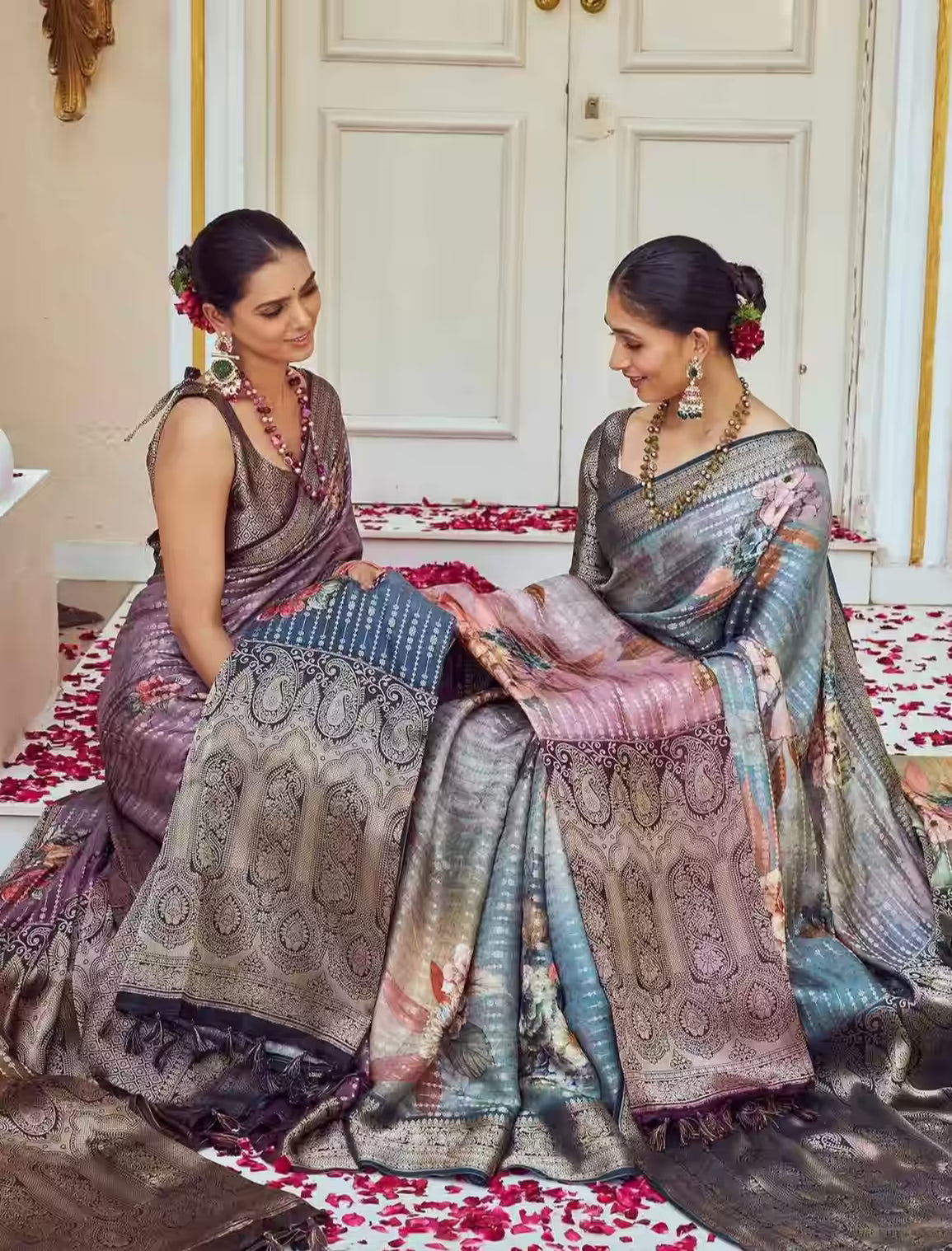 Two women in traditional sarees sitting on a decorated floor with floral arrangements.